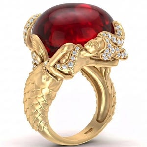 May include: A gold ring featuring a large, oval, deep red cabochon gemstone held by two sculpted figures with textured, scale-like lower bodies. The figures are adorned with small, clear, sparkling stones along their hair and arms.