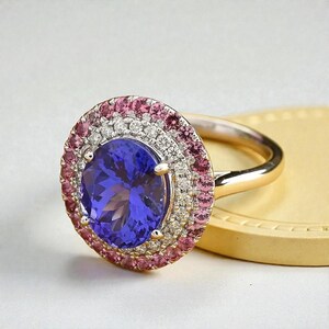May include: A gold ring featuring a large, oval-cut, deep blue gemstone surrounded by a halo of smaller pink and white gemstones. The ring's band is a polished gold color, and the setting is intricate.
