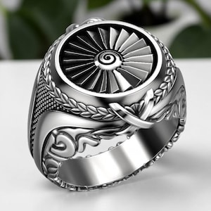 May include: A silver-colored ring with a detailed design. The ring features a central turbine-like element, surrounded by intricate patterns and a laurel wreath design. The ring has a polished, metallic finish.