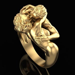 May include: A gold-toned ring featuring a sculptural design of two figures in an embrace. The figures are depicted with detailed hair and limbs, forming the band of the ring. The surface has a polished, metallic finish with smooth contours.