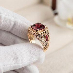 Può includere: Un anello in oro con una grande pietra preziosa rosso scuro a taglio quadrato al centro, affiancata da pietre rosse più piccole a taglio marquise e un intricato lavoro di filigrana metallica lungo la fascia, con dettagliati motivi a volute.