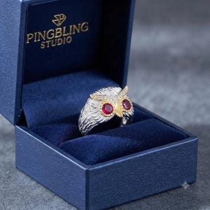 Peut inclure: Une bague hibou argentée avec deux yeux en pierre rouge ronde et un bec doré, placée dans un écrin bleu marine avec le texte PINGBLING STUDIO imprimé en doré sur le couvercle intérieur. L'anneau présente un motif de plumes texturé.