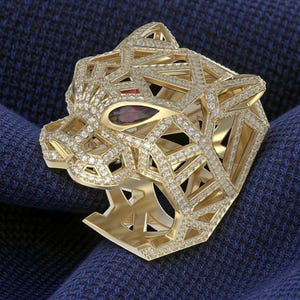 May include: A gold-colored ring in the shape of a panther's head, encrusted with small, sparkling diamonds. The panther has a red gemstone eye. The ring is set against a dark blue textured background.