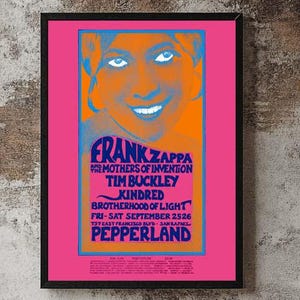 Puede incluir: Un póster enmarcado con un vibrante fondo rosa presenta un retrato de Frank Zappa en naranja y azul. El póster incluye el texto "FRANK ZAPPA" y otros nombres de bandas. Un tocadiscos negro está sobre un mueble de madera.
