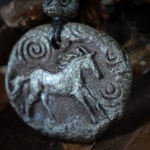 May include: A circular dark gray pendant with a textured, stone-like finish featuring a relief carving of a horse in motion. The pendant includes a spiral design and crescent shapes, suspended from a black cord with a matching dark gray bead.