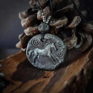 May include: A circular dark gray pendant featuring a raised silver-toned horse relief, spiral and crescent moon symbols, suspended on a black cord with small metallic beads. The textured, coin-like piece displays a weathered, metallic finish.