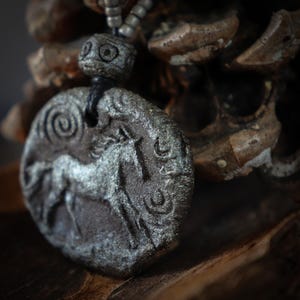 May include: A circular dark gray pendant featuring a raised relief of a horse in motion, accented with a spiral and crescent moon symbols. The pendant hangs from a dark cord adorned with small, light-colored cylindrical beads above the main piece.