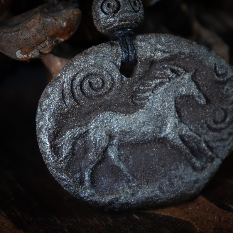 May include: A dark gray, oval-shaped pendant featuring a relief carving of a horse in motion. The textured surface includes spiral patterns and is suspended by a black cord through a hole at the top, accompanied by a matching dark gray bead.