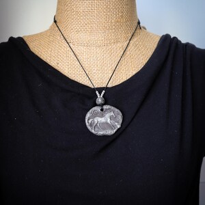 May include: A silver-toned circular pendant featuring a relief of a horse in motion, suspended from a thin black cord. The pendant has a textured, weathered surface and is accented with small silver beads above the main circular charm.