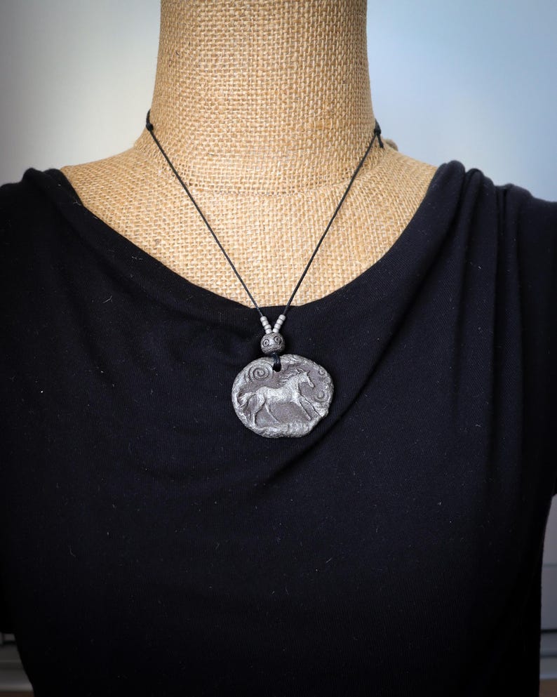 May include: A silver-toned circular pendant featuring a relief of a horse in motion, suspended from a thin black cord. The pendant has a textured, weathered surface and is accented with small silver beads above the main circular charm.