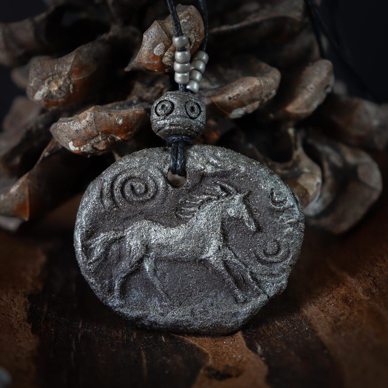 May include: A dark gray, textured, oval-shaped pendant featuring a raised relief of a horse in motion. The pendant is suspended from a black cord adorned with small, metallic, cylindrical beads and a larger, round, textured bead above the main piece.