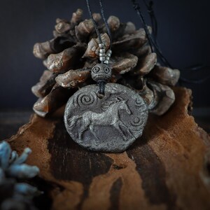 May include: A circular, textured gray pendant featuring a relief of a galloping horse with a spiral and crescent moon symbol. The pendant hangs from a black cord with small silver-toned beads and a decorative, etched spherical bead above it.