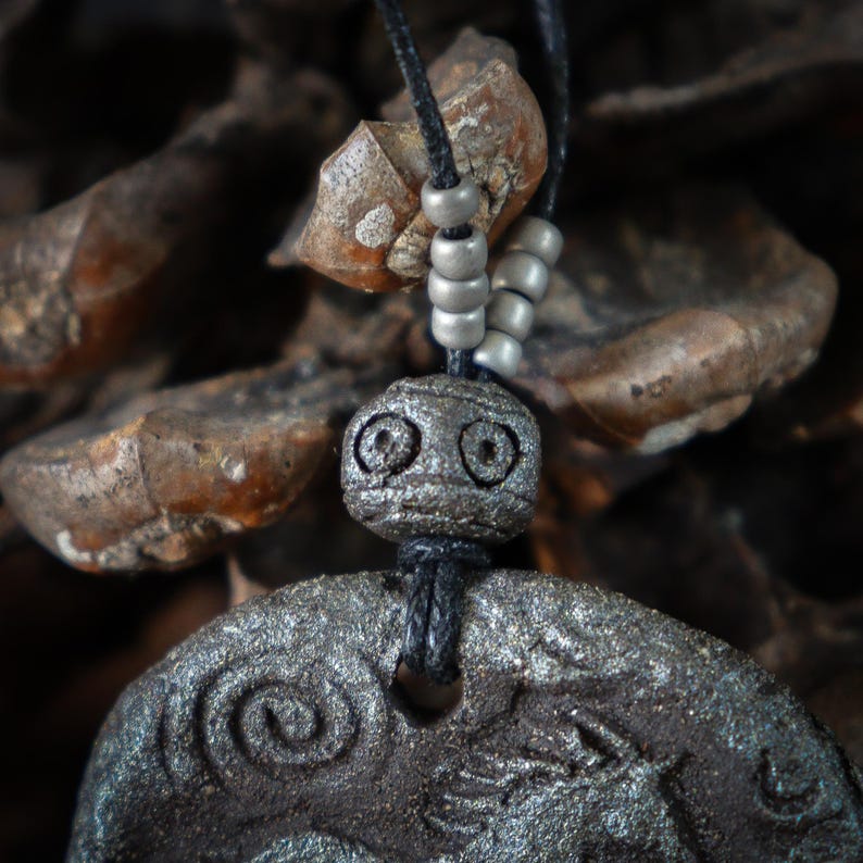 May include: A dark gray, textured circular pendant featuring an embossed spiral and animal figure, suspended from a black cord. The cord is accented with four small, matte silver-toned cylindrical beads and a single larger, carved spherical bead.
