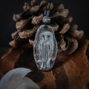 May include: A dark brown, oval-shaped clay pendant featuring a textured, white-washed relief of an owl. The pendant hangs from a black cord adorned with small, dark, spherical beads, creating a contrast between the dark base and the light owl.