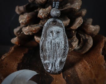 May include: A dark brown, oval-shaped clay pendant featuring a textured, white-washed relief of an owl. The pendant hangs from a black cord adorned with small, dark, spherical beads, creating a contrast between the dark base and the light owl.