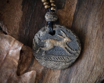May include: A circular dark gray clay pendant featuring a gold-toned relief of a running hare above textured ground. The pendant hangs from a black cord adorned with three small metallic beads and one larger textured bead above the main piece.