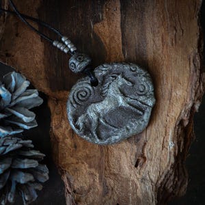 May include: A circular gray pendant featuring a relief carving of a horse and spiral patterns. The pendant is suspended from a black cord adorned with small gray beads. The textured surface of the pendant has a weathered, stone-like appearance.