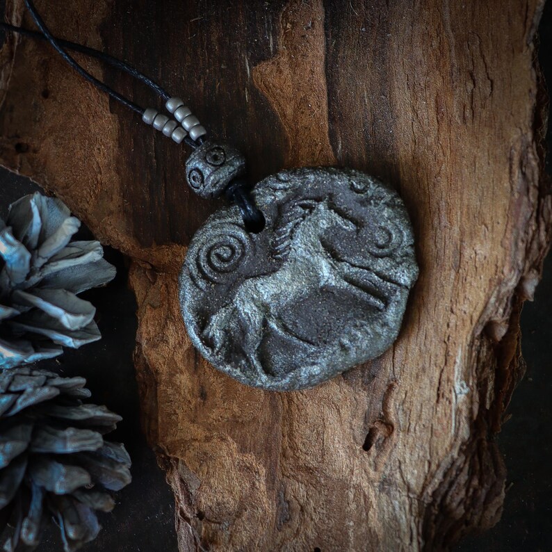 May include: A circular gray pendant featuring a relief carving of a horse and spiral patterns. The pendant is suspended from a black cord adorned with small gray beads. The textured surface of the pendant has a weathered, stone-like appearance.