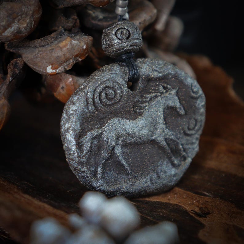 May include: A dark gray, textured, oval-shaped pendant featuring a relief carving of a horse in motion. The pendant is suspended from a black cord with a matching dark gray, cylindrical, patterned bead above the main piece.
