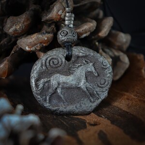 May include: A dark gray, textured, oval-shaped pendant featuring a relief carving of a horse in motion. The pendant is suspended from a black cord adorned with small, metallic, cylindrical beads and a larger, round, etched bead above the main piece.