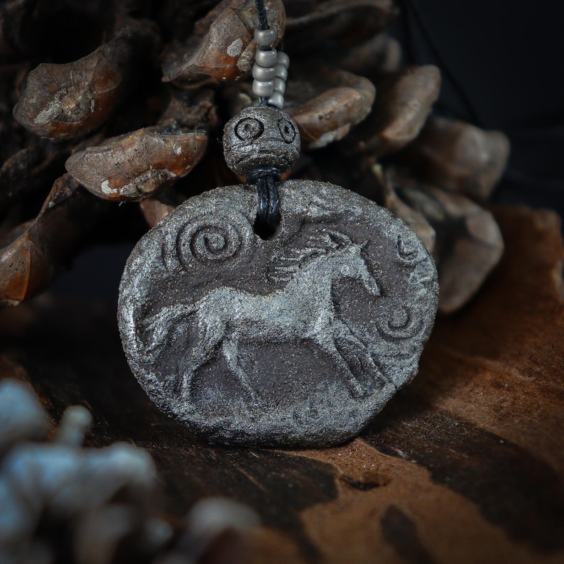 May include: A dark gray, textured, oval-shaped pendant featuring a relief carving of a horse in motion. The pendant is suspended from a black cord adorned with small, metallic, cylindrical beads and a larger, round, etched bead above the main piece.