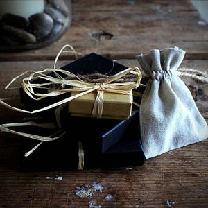 May include: A stack of three square black gift boxes topped with a smaller tan cardboard box, all secured with natural raffia ribbon. Beside the boxes sits a small, light beige drawstring pouch made of woven fabric with a jute cord tie.