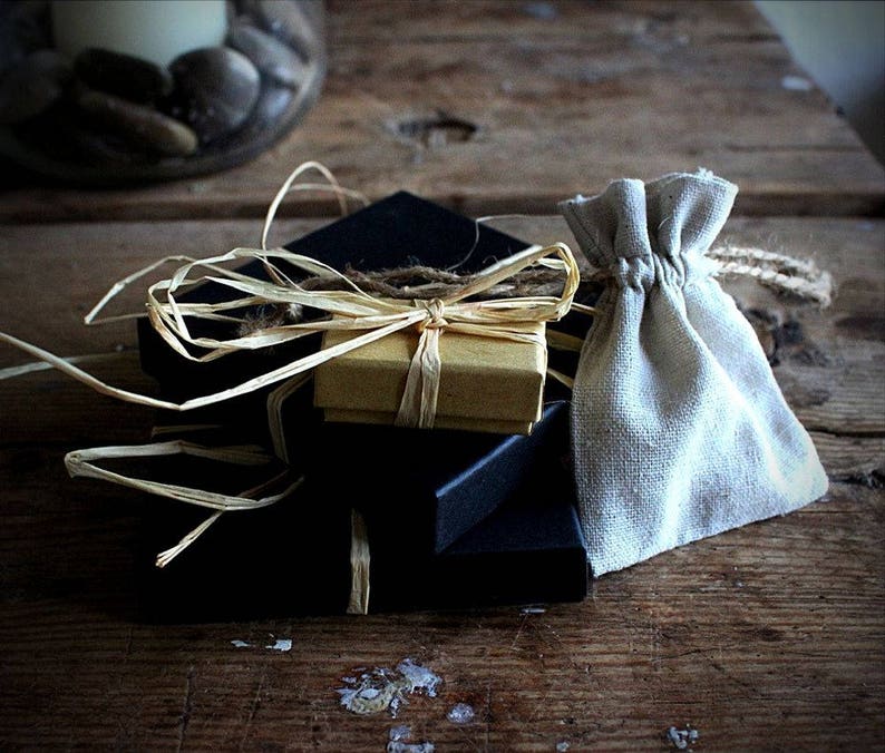 May include: A stack of three square black gift boxes topped with a smaller tan cardboard box, all secured with natural raffia ribbon. Beside the boxes sits a small, light beige drawstring pouch made of woven fabric with a jute cord tie.