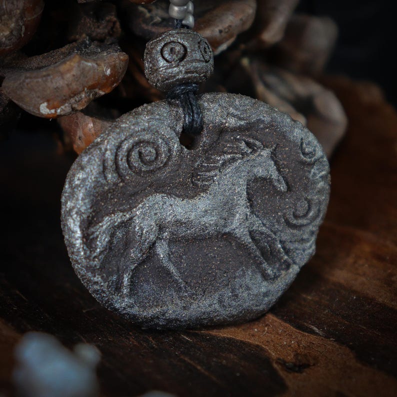 May include: A dark gray, textured, oval-shaped pendant featuring a relief carving of a horse in motion. The pendant is suspended by a black cord with a matching dark gray, spherical, patterned bead positioned directly above the main piece.