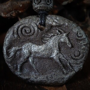 May include: A dark gray, oval-shaped pendant featuring a textured, embossed horse figure in the center. The surface has a metallic, shimmering finish with spiral patterns on the sides. A black cord passes through a hole at the top, secured by a bead.