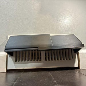 Op de afbeelding: Een zwarte metalen ventilatieafdekking met een rechthoekig ontwerp. De ventilatieafdekking is geïnstalleerd op een witte plint en staat op een donkerbruine houten vloer. De muur achter de ventilatie is lichtgrijs geschilderd.