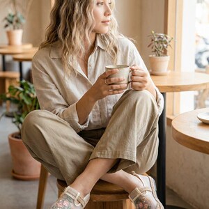 May include: A person wearing a beige linen shirt and trousers, seated on a wooden stool, holding a mug. They are wearing floral embroidered espadrille shoes. The scene is set in a cafe with round tables and potted plants.
