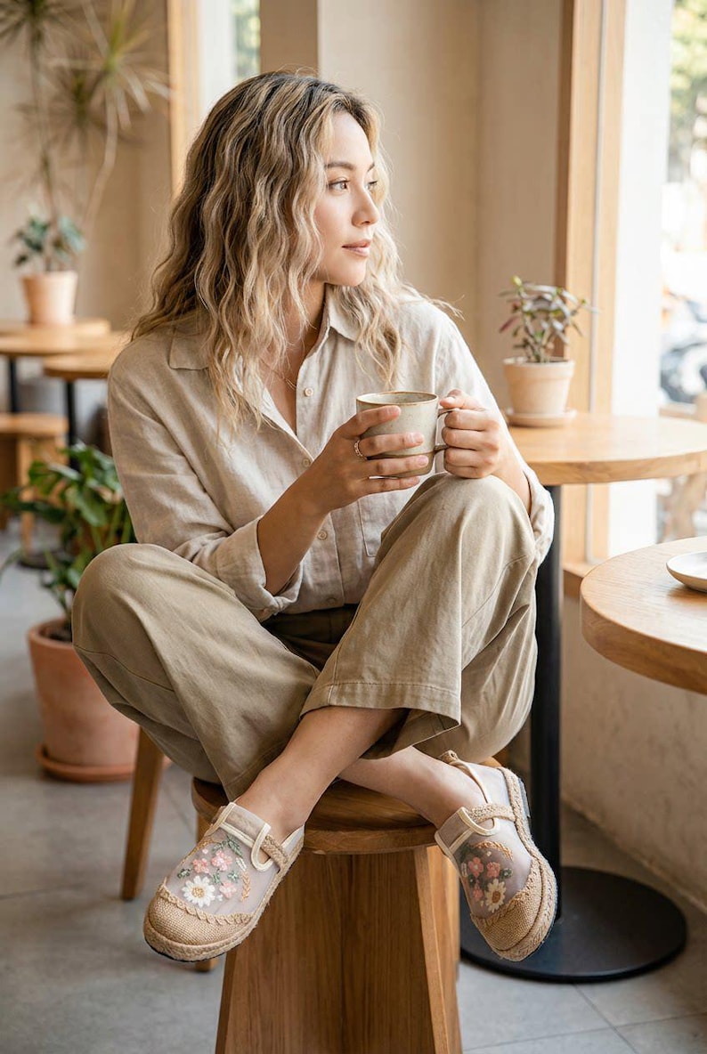 May include: A person wearing a beige linen shirt and trousers, seated on a wooden stool, holding a mug. They are wearing floral embroidered espadrille shoes. The scene is set in a cafe with round tables and potted plants.