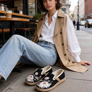 K&ouml;nnte beinhalten: Schwarze Espadrilles zum Hineinschl&uuml;pfen mit floraler Stickerei. Die Schuhe haben ein Obermaterial aus schwarzem Canvas, eine Sohle aus nat&uuml;rlichem Juteseil und eine wei&szlig;e Einfassung. Das florale Design zeigt rosa und wei&szlig;e Blumen mit gr&uuml;nen Bl&auml;ttern.