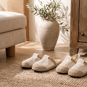 Può includere: Due paia di scarpe slip-on in pizzo bianco e un paio di pantofole espadrillas in pizzo bianco con suole in juta sono disposte su un tappeto tessuto. Un divano beige e un armadio in legno sono sullo sfondo.
