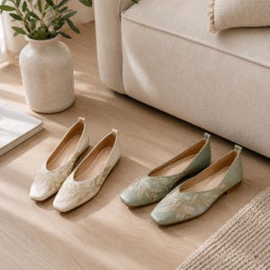 May include: Two pairs of embroidered flats in ivory and sage green. The ivory flats have gold floral embroidery, while the sage green flats feature gold leaf embroidery. The shoes are displayed on a light wood floor near a beige sofa.