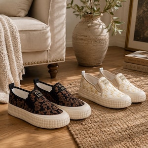 May include: Two pairs of slip-on trainers. One pair features a black floral pattern with a white sole, and the other pair has a cream floral design with a white sole. Both pairs have a small logo on the top.