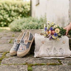 Puede incluir: Un par de zapatos espadrilles de lona gris con diseños florales bordados, junto a un delantal de lino beige con bordados florales y un ramo de flores silvestres coloridas. Los artículos están dispuestos sobre una superficie de piedra.