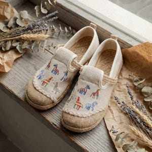 Può includere: Un paio di scarpe slip-on beige con suola in iuta e tomaia in tela. La tela è ricamata con disegni di cavalli colorati. Le scarpe sono esposte su una superficie di legno con piante essiccate.
