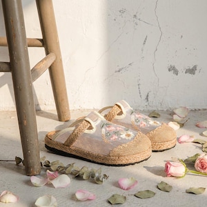 May include: A pair of beige and tan embroidered floral slippers with a woven jute sole. The slippers feature a sheer overlay with embroidered flowers and a braided strap. The background includes a wooden stool and scattered rose petals.