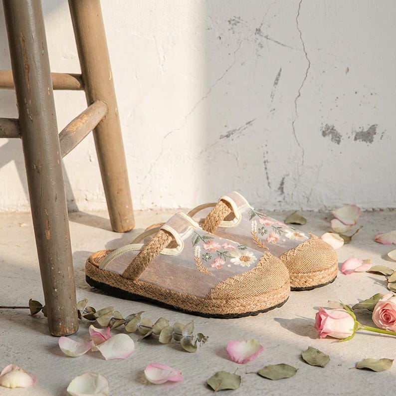 May include: A pair of beige and tan embroidered floral slippers with a woven jute sole. The slippers feature a sheer overlay with embroidered flowers and a braided strap. The background includes a wooden stool and scattered rose petals.