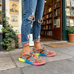 Puede incluir: Zapatos sin cordones multicolores con un vibrante estampado psicodélico. Los zapatos presentan una suela degradada arcoíris y una mezcla de diseños en zigzag y ondulados en tonos rosa, amarillo, azul y verde. Llevados con vaqueros azules desgastados.