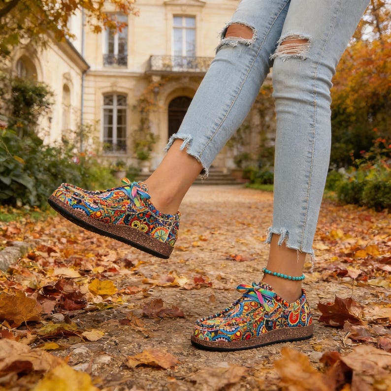 May include: Brightly patterned slip-on shoes with a cork sole. The shoes display a vibrant mix of colours and designs, including blue, orange, and yellow. The shoes are shown with light blue ripped jeans and a turquoise bracelet.