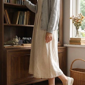Pode incluir: Uma mulher com um vestido bege claro e um cardigan cinzento está perto de uma estante de madeira. Ela usa espadrilles bege. A sala tem uma câmara vintage, flores secas e um cesto de vime.
