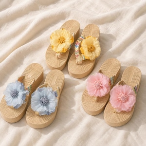 Op de afbeelding: Vier plateau-slippers met geweven beige zolen en veelkleurige chevronbandjes. Elk paar heeft een grote, doorschijnende stoffen bloem in geel, lichtblauw of roze, bevestigd in het midden van het bandje, wat zorgt voor een textuurrijk bloemendesign.