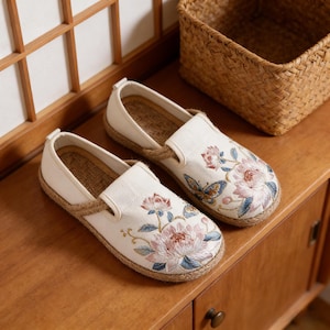 K&ouml;nnte beinhalten: Ein Paar cremefarbene Slipper mit floraler Stickerei. Die Schuhe haben eine Sohle aus Juteseil und sind mit rosa, blauen und gr&uuml;nen gestickten Blumen und Schmetterlingen verziert. Die Schuhe werden auf einer Holzoberfl&auml;che pr&auml;sentiert.
