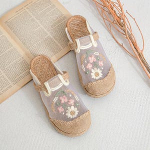 May include: Embroidered floral slippers. The slippers have a woven jute sole, a sheer lavender upper with pink and white floral embroidery, and a woven strap. The design includes a beige trim.