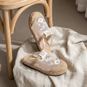 May include: A pair of beige espadrille sandals with floral embroidery. The sandals have a woven jute sole, sheer fabric overlay, and delicate pink and white flower embroidery. The straps are a light beige colour.