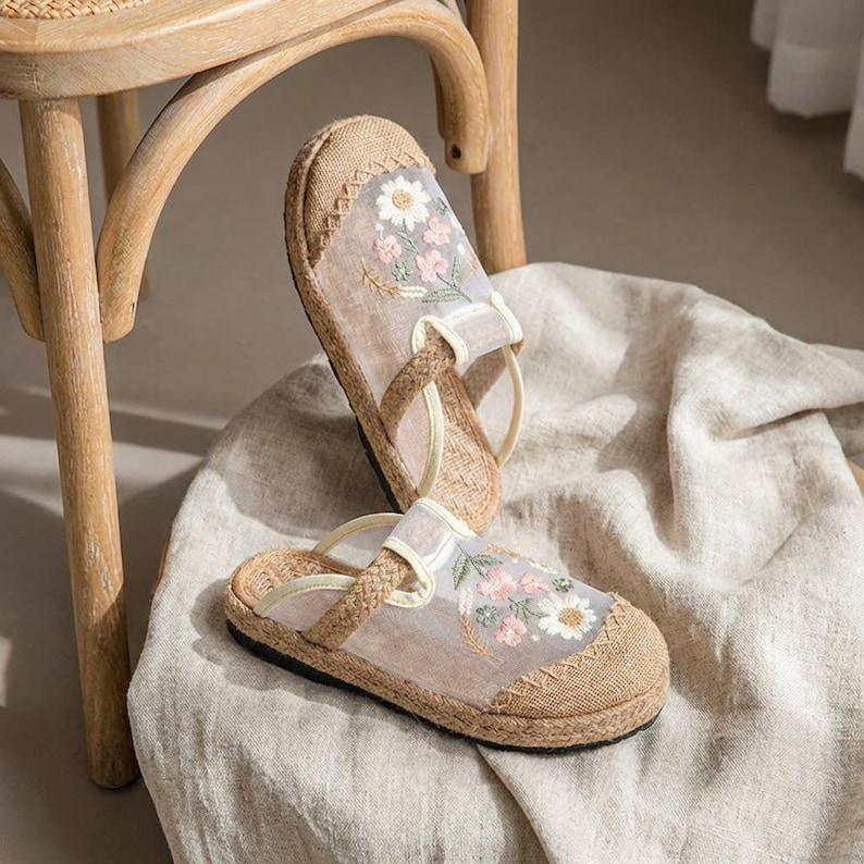 May include: A pair of beige espadrille sandals with floral embroidery. The sandals have a woven jute sole, sheer fabric overlay, and delicate pink and white flower embroidery. The straps are a light beige colour.