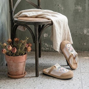 May include: Beige and white floral sandals with jute soles sit on a speckled floor. A vintage wooden chair with a woven seat and a beige cloth draped over it is in the background. A terracotta pot with dried roses is also visible.