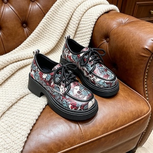 Könnte beinhalten: Schwarze Plateauschuhe mit floralem und Totenkopf-Muster in Rot, Türkis und Weiß. Die Schuhe haben schwarze Schnürsenkel und eine dicke schwarze Sohle. Die Schuhe werden auf einem braunen Ledersessel präsentiert.